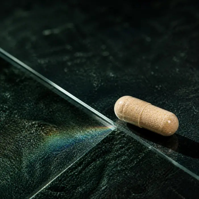 One capsule resting on dark green stone beside the edge of a glass prism casting a rainbow light refraction
